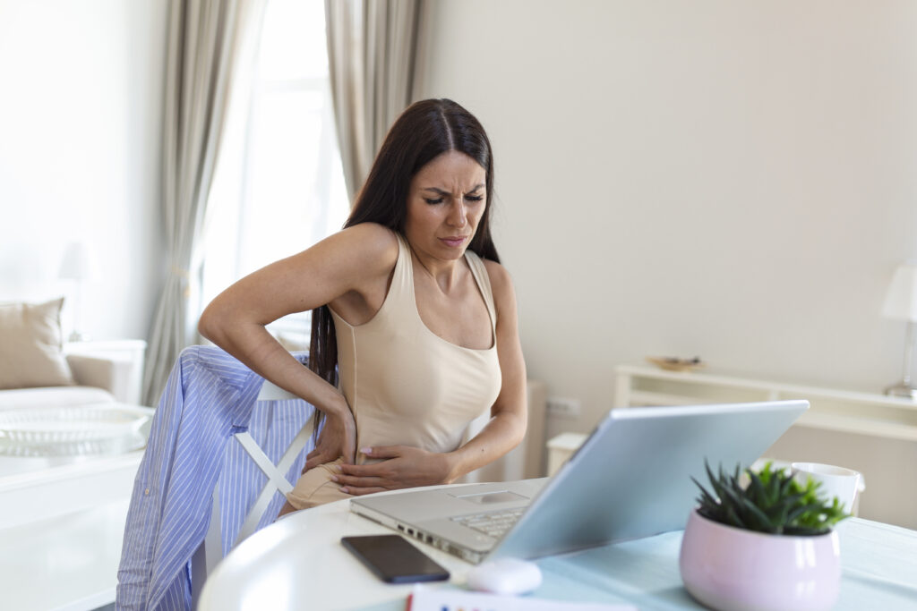 Businesswoman Holding Her Back While Working On Laptop At Home Office Desk. young woman, stretching, sitting feeling her hip pain tired after working at laptop, small home office interior.