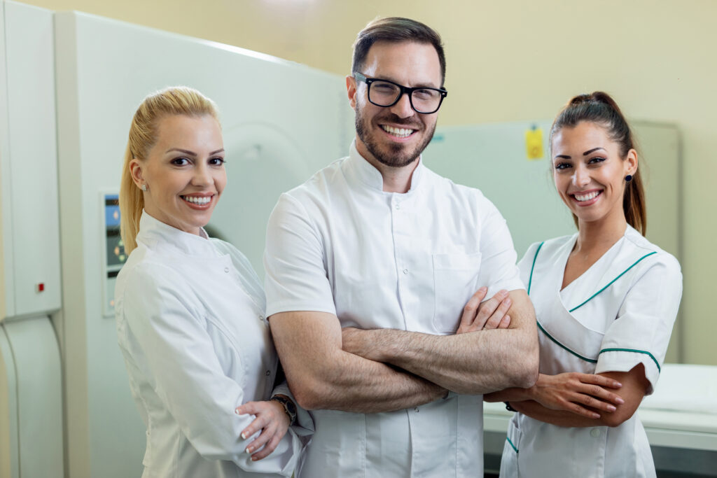 Team of happy doctors standing with arms crossed next to MRI scanner in the hospital and looking at camera. About.